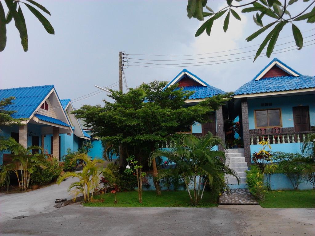 Two colorful bungalows with blue roofs are visible, surrounded by lush greenery and small plants. A pathway leads to the entrance of each unit, framed by neatly maintained lawns. Nearby utility poles are present against a gray sky, adding a touch of ambiance to the scene.