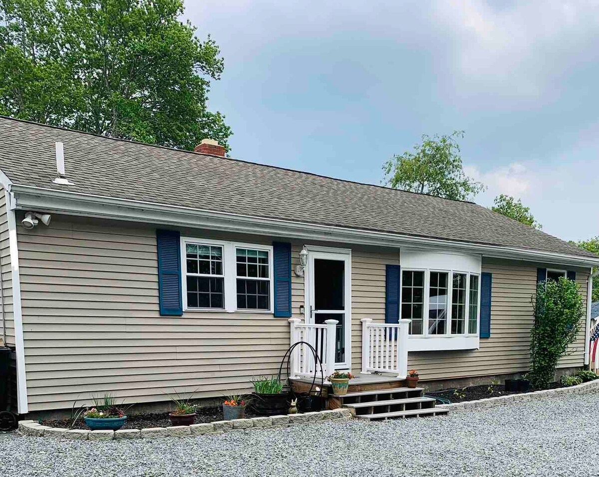 The exterior of the lakeside home is shown, featuring a beige facade with blue shutters and a welcoming front porch. A gravel pathway leads to the entrance, surrounded by flower beds. Trees provide greenery in the background, contributing to the tranquil setting.