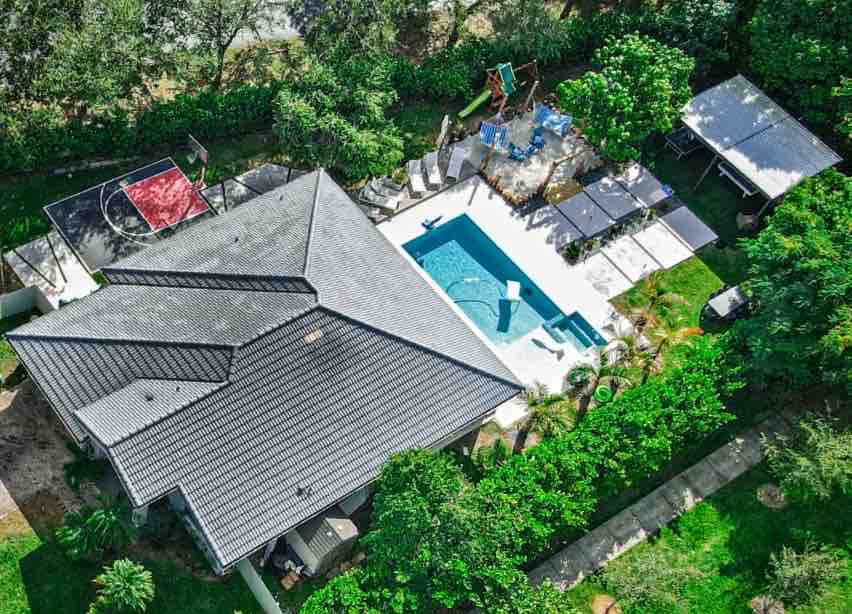 An aerial view captures the modern home with a landscaped yard, featuring a heated pool surrounded by sunbeds and shaded cabanas. The outdoor play area includes a basketball hoop and playground equipment, all framed by lush greenery for added privacy.