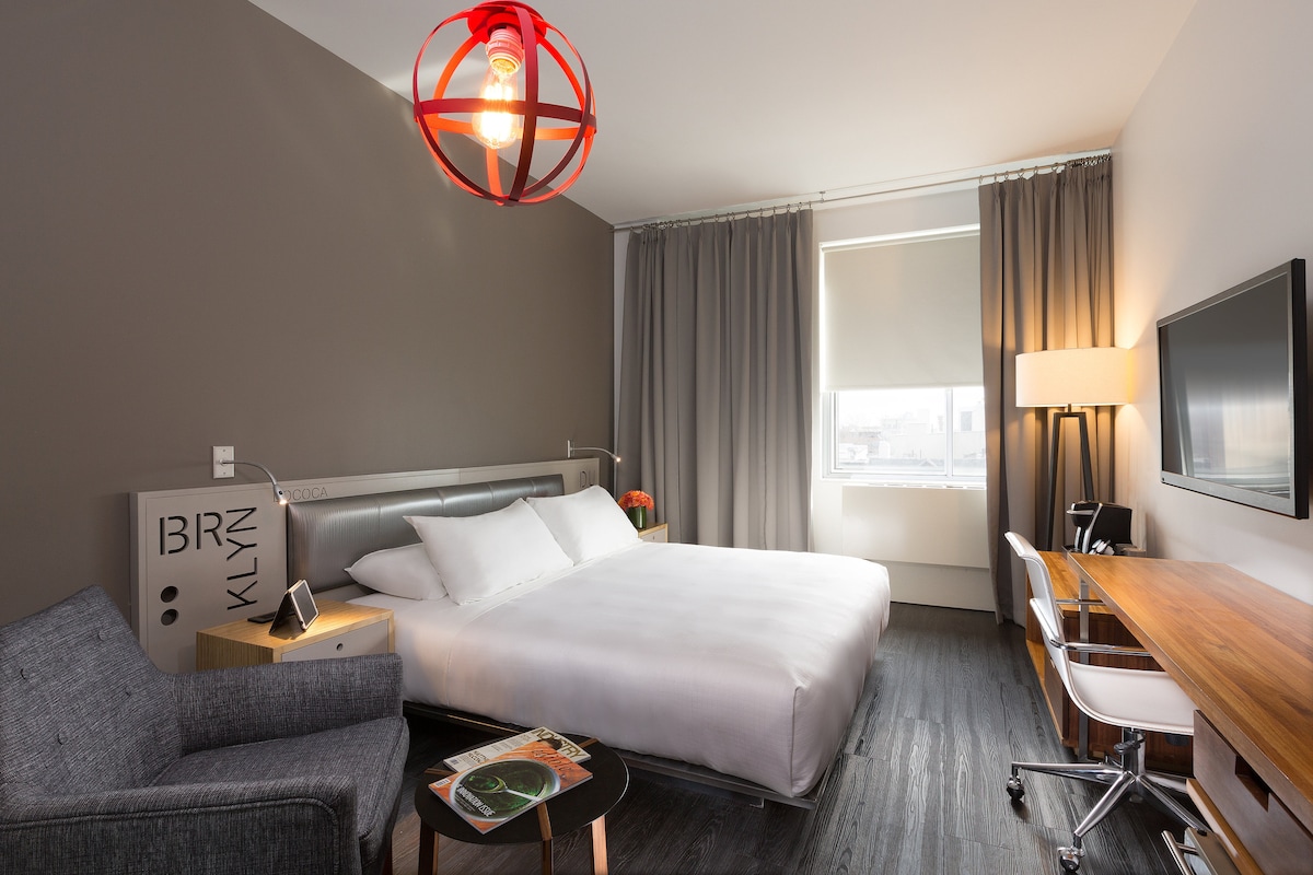 A modern room is furnished with a spacious bed and a wooden desk. A comfortable chair sits beside a small table, while a flat-screen TV is mounted on the wall. Natural light filters through layered curtains, complemented by contemporary lighting fixtures.