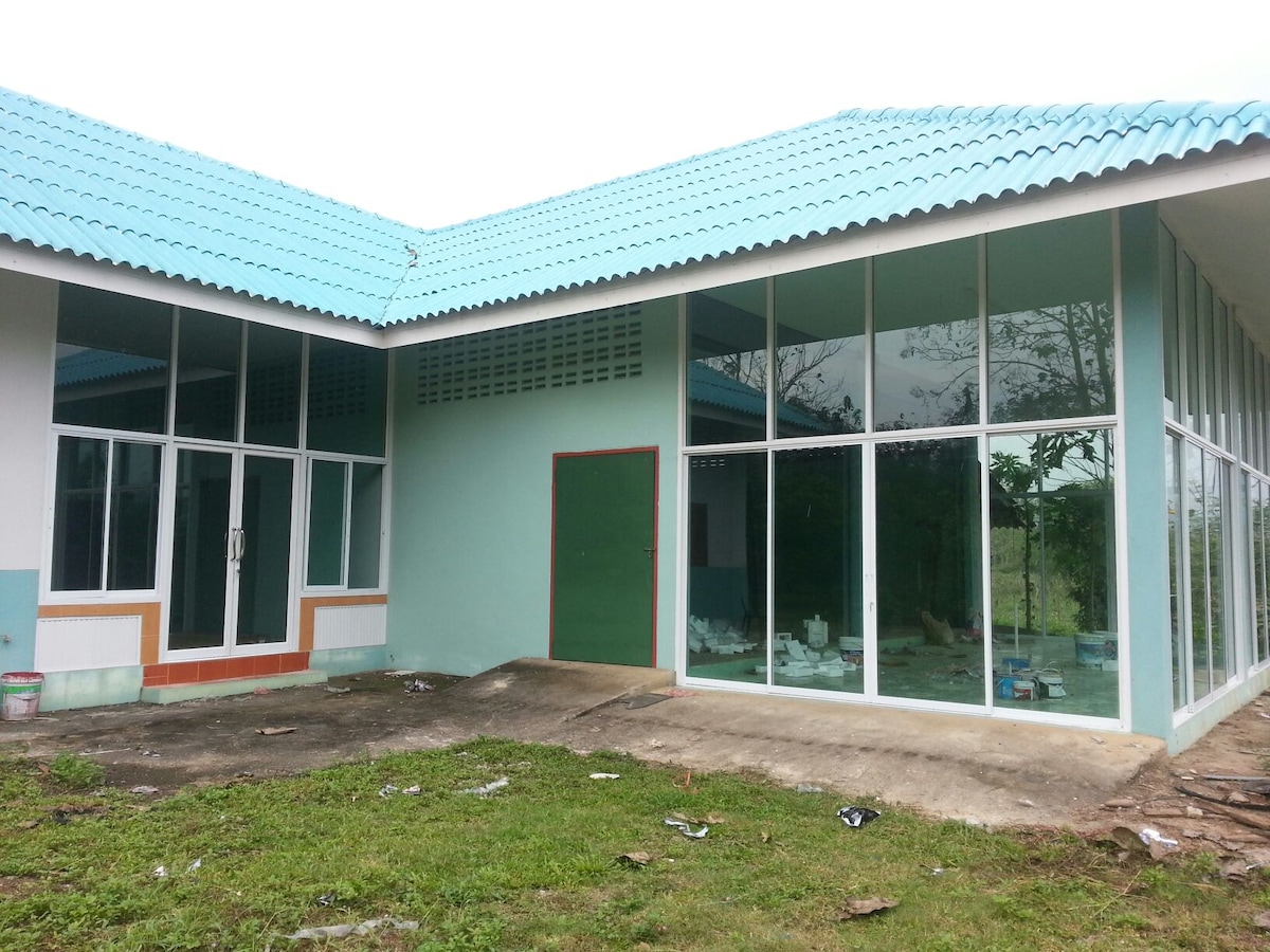The exterior of the country home is showcased with large glass windows and a green door, allowing natural light to brighten the interior. The low-pitched roof is covered with blue tiles, and the grass surrounding the structure appears well-maintained.