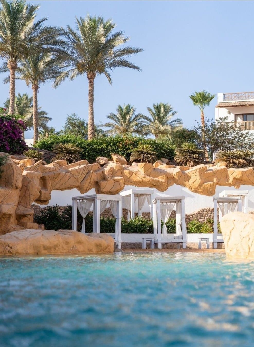 A serene outdoor lounging area is framed by lush palm trees and natural rock formations. White cabanas with sheer curtains are placed over a smooth stone surface, offering shaded spots for relaxation beside a crystal-clear pool.