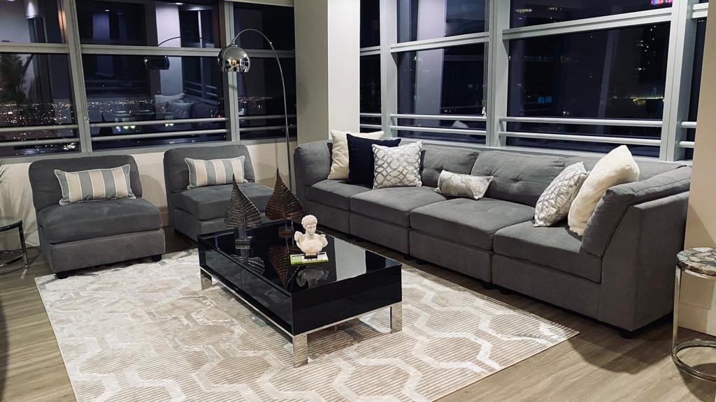 A modern living area features a gray sectional sofa complemented by a matching accent chair. A sleek black coffee table sits atop a light rug, with decorative sculptures arranged thoughtfully. Large windows provide expansive views of the city skyline, allowing natural light to fill the space.