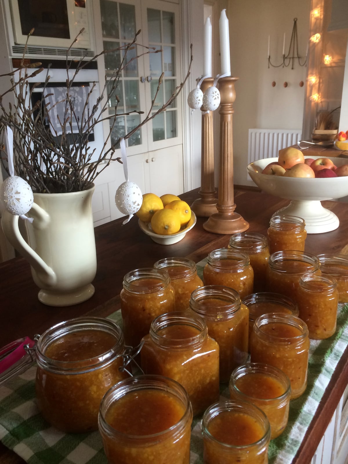A wooden table is adorned with several jars of homemade preserves, varying in size, and arranged in a neat row. Decorative eggs hang from branches in a vase, while a bowl filled with lemons and a fruit bowl hold fresh apples. Subtle lighting enhances the welcoming atmosphere.