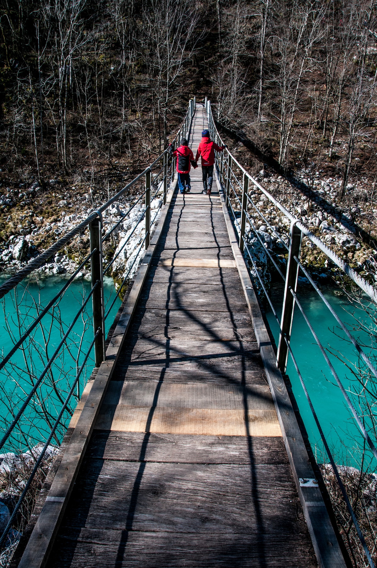 A narrow wooden suspension bridge spans a vibrant turquoise river, flanked by rocky banks. Two individuals in red jackets walk side by side, casting elongated shadows on the bridge's surface, surrounded by trees with bare branches.