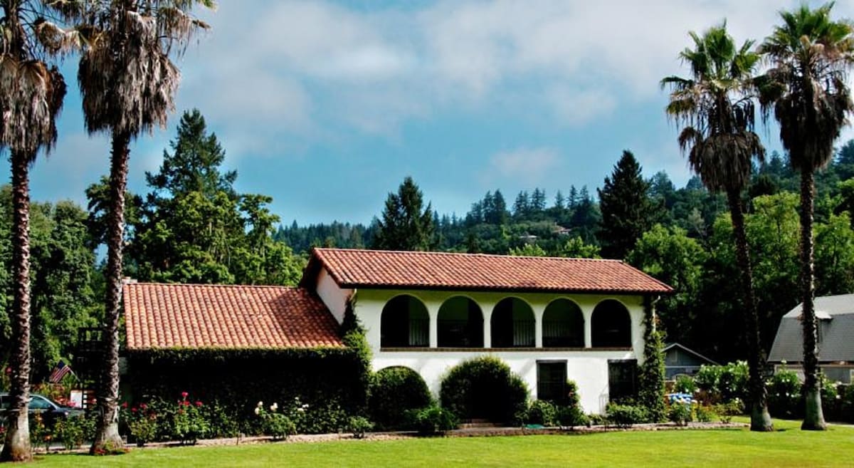 The Mediterranean villa is framed by tall palm trees and lush greenery. The facade features a red tiled roof and arched windows, creating a harmonious blend with the surrounding landscape. A well-maintained lawn stretches in front, enhancing the villa's inviting presence.