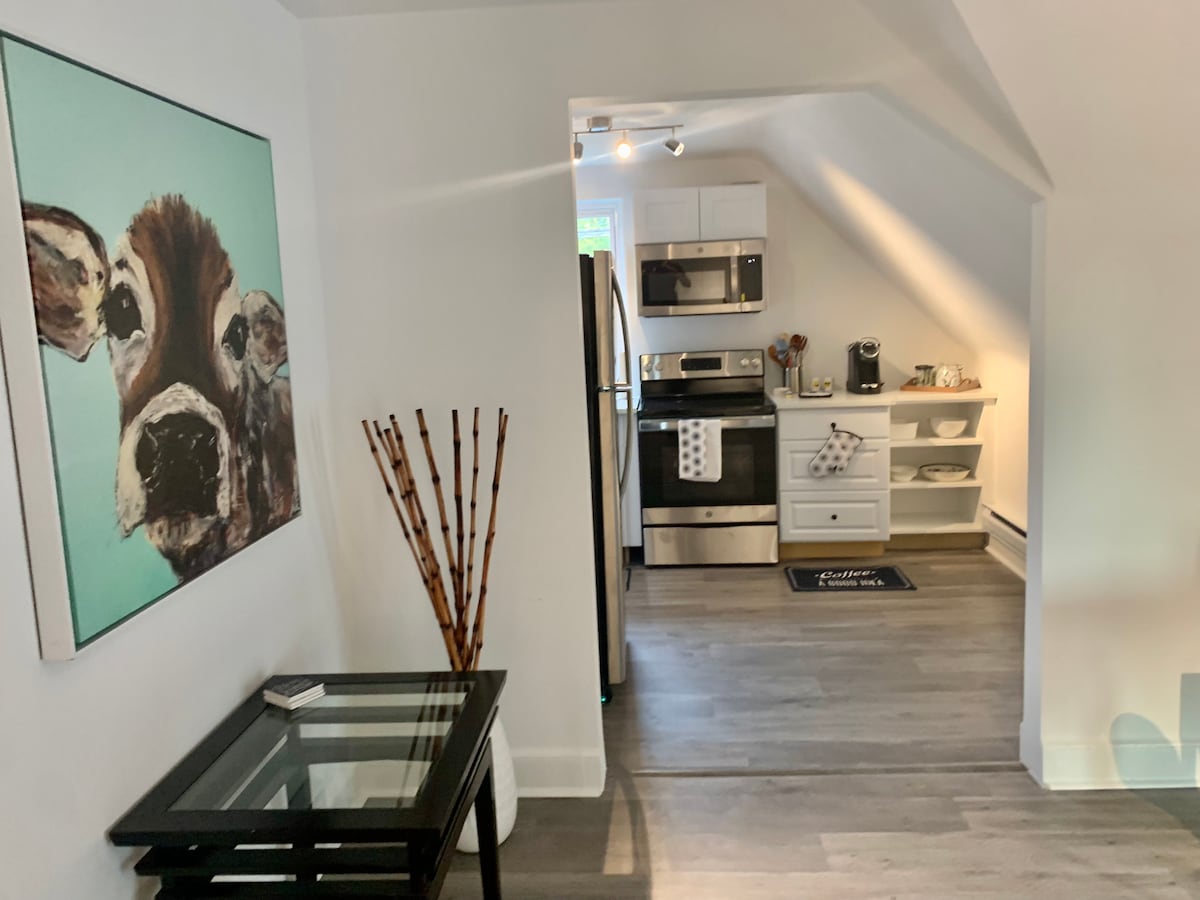 The kitchen area is visible, featuring modern stainless steel appliances and a well-organized countertop. A painted artwork of a cow adorns the wall, accompanied by decorative wooden sticks in a vase. Natural light fills the space, highlighting the open layout.