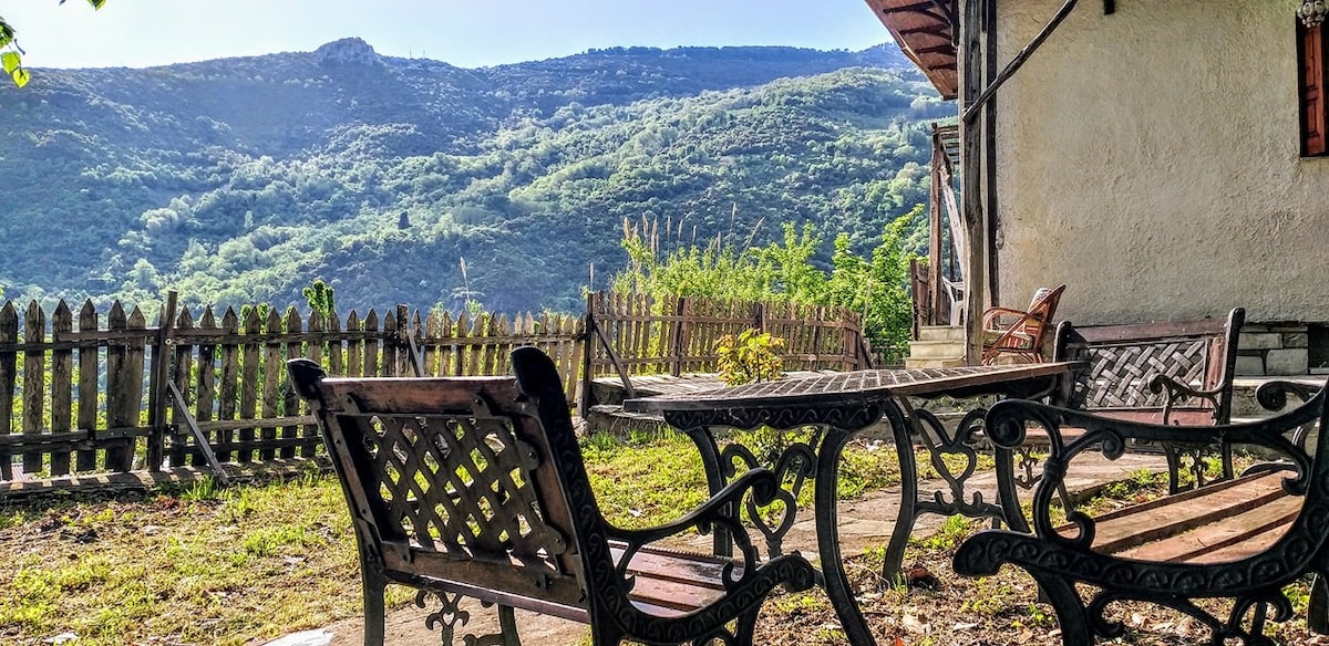 An outdoor seating area is displayed with wooden tables and chairs arranged on a grassy patch. A wooden fence provides a rustic border, with mountains and lush greenery visible in the background under a clear sky.