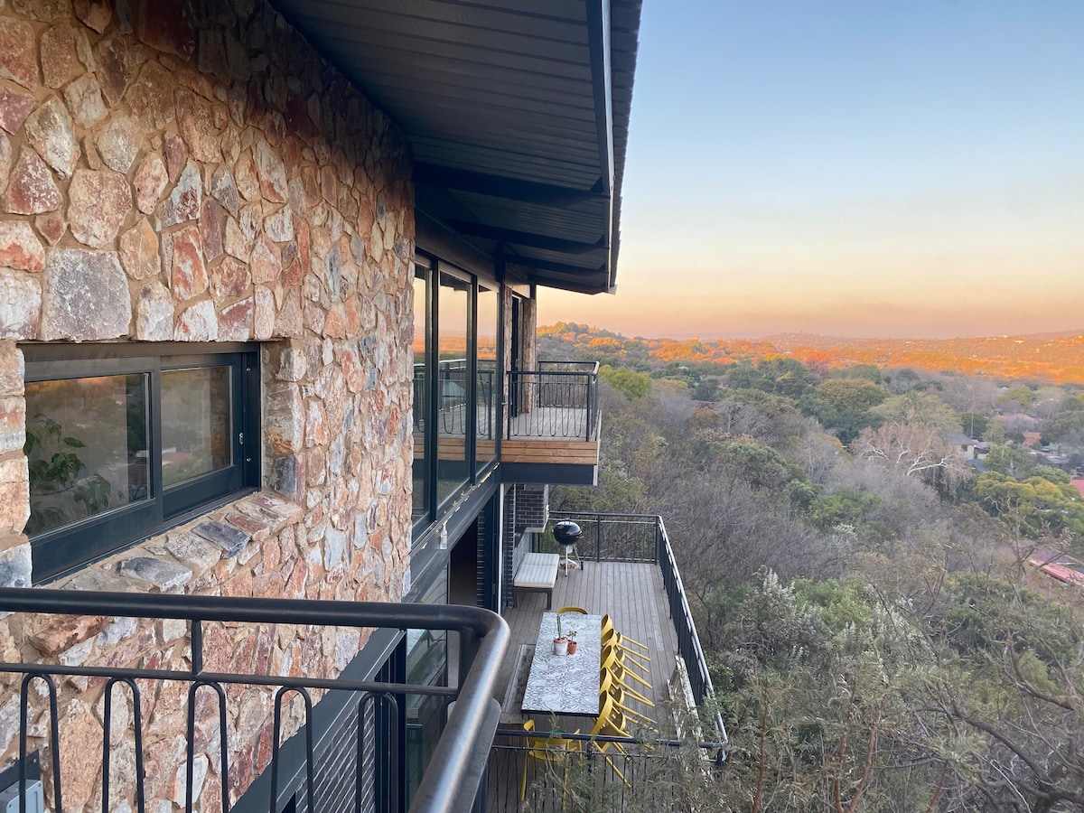 A balcony view showcases a stone and steel structure nestled among jacaranda treetops, with expansive views of the city skyline. A table with yellow chairs is positioned along the balcony, inviting relaxation in the serene outdoor setting.