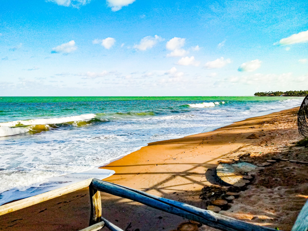 An expansive view of a sandy beach features gentle waves lapping against the shore under a clear blue sky with scattered clouds. The shoreline is outlined by a wooden railing, indicating a welcoming access point to the beach.