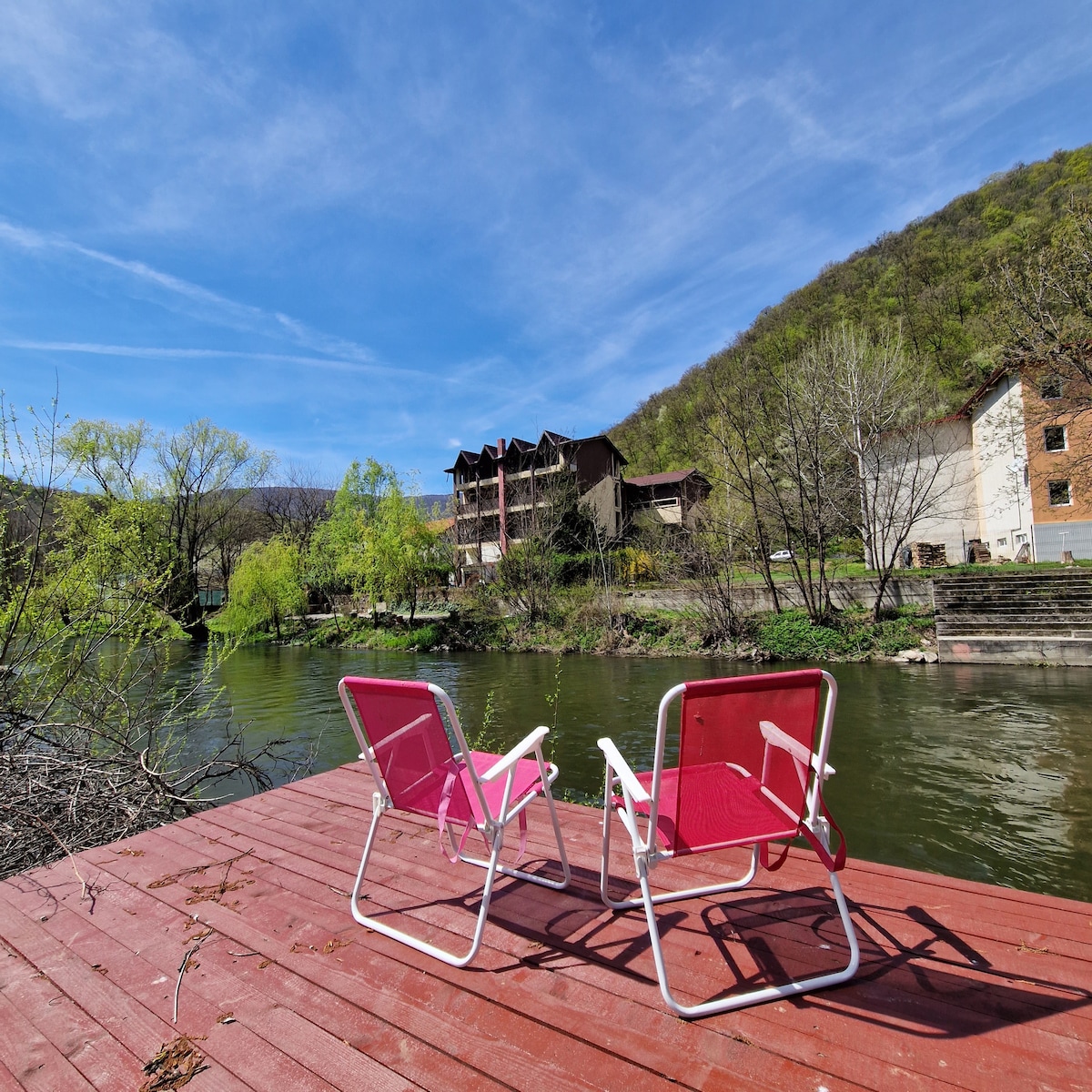 Two bright pink chairs are positioned on a wooden dock extending over a calm river. Lush greenery and trees line the banks, while a multi-story building is visible in the background, surrounded by hillside vegetation. A clear blue sky enhances the serene setting.