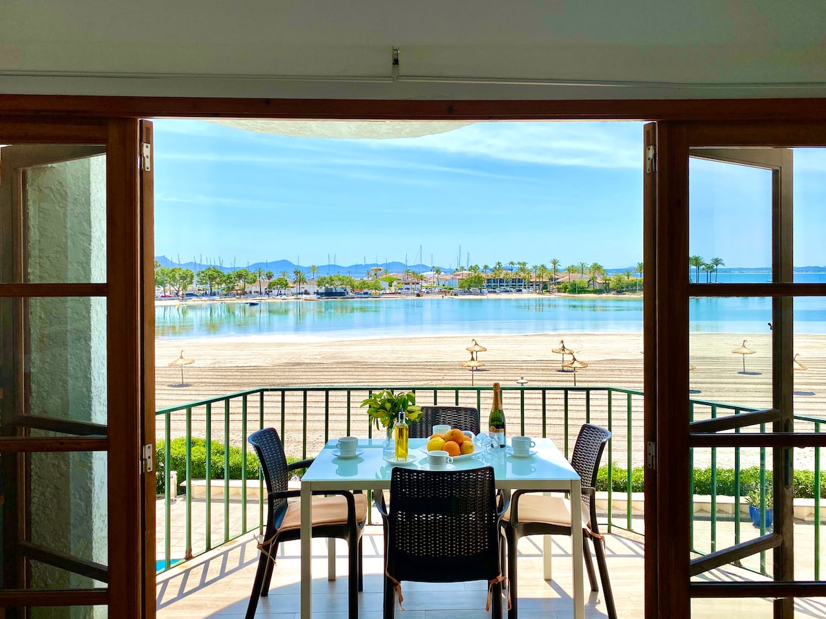 A balcony offers a panoramic view of the sandy beach and calm waters, framed by open wooden doors. A dining table is set for four, featuring fresh fruit and a vase with flowers. The serene landscape in the background includes palm trees and distant mountains.