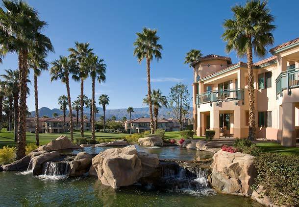 A serene landscape features a cascading water feature surrounded by smooth stones, vibrant greenery, and palm trees. The two-story villas are visible in the background, with balconies overlooking the tranquil scenery and distant mountains under a clear blue sky.
