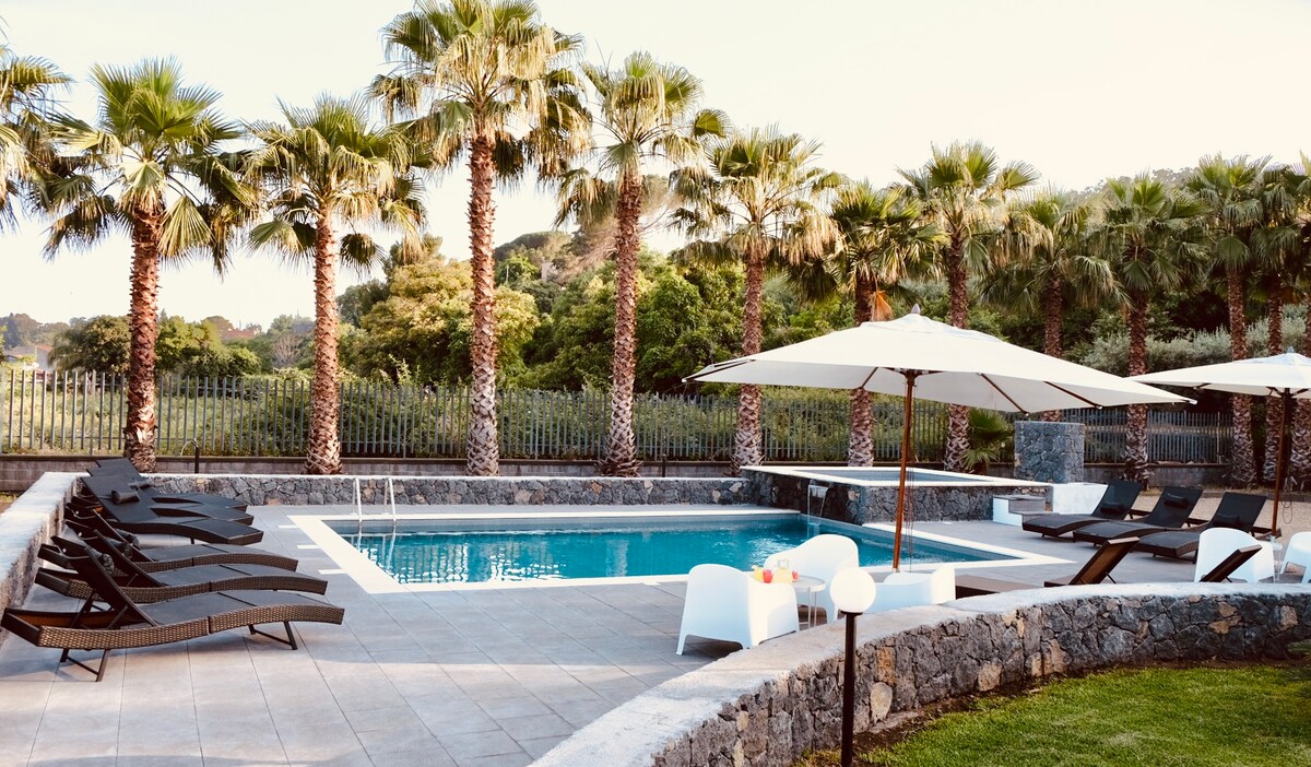 A swimming pool is framed by tall palm trees, providing a relaxing atmosphere. Sun loungers are positioned around the pool, and umbrellas offer shaded seating. Natural stone and tiled surfaces create a seamless connection to the surrounding greenery.
