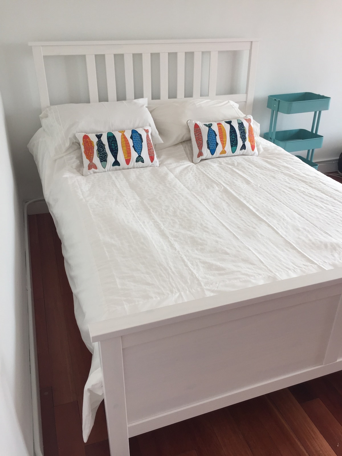 A spacious queen bed is neatly made with white linens, complemented by two decorative pillows featuring colorful fish prints. A small, teal nightstand is positioned beside the bed, contributing to a fresh and airy feel.