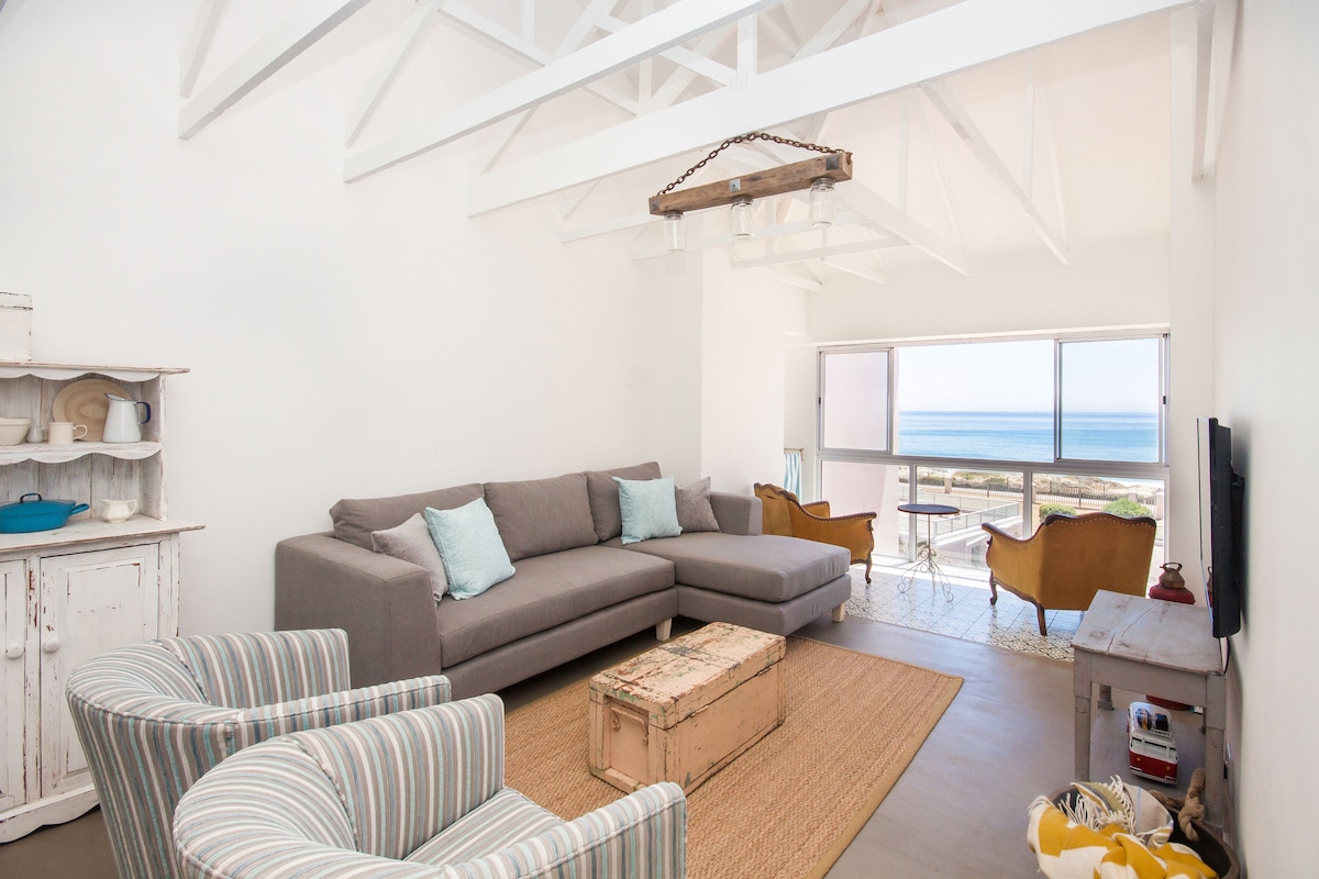 A light-filled living area features a spacious gray sectional sofa and two armchairs with a coastal view. A rustic coffee table sits in front of the sofa, while a large window invites the ocean scenery and natural light into the space.
