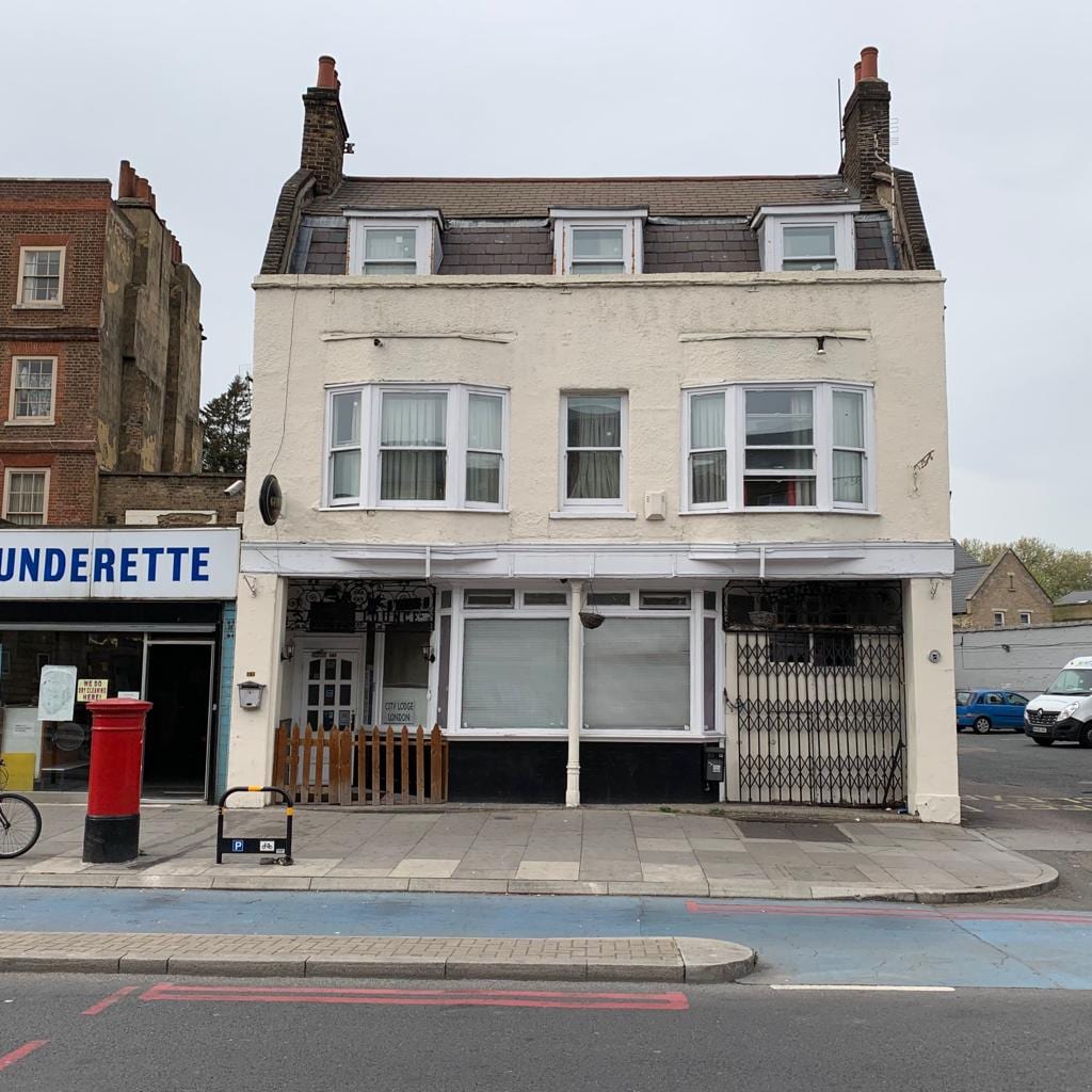 A three-story building is visible with a white exterior and a sloped roof. Large windows are featured on the upper floors, and a ground-level entrance is presented. A shop front, labeled 'UNDERETTE,' is situated to the left, with a red post box in the foreground.