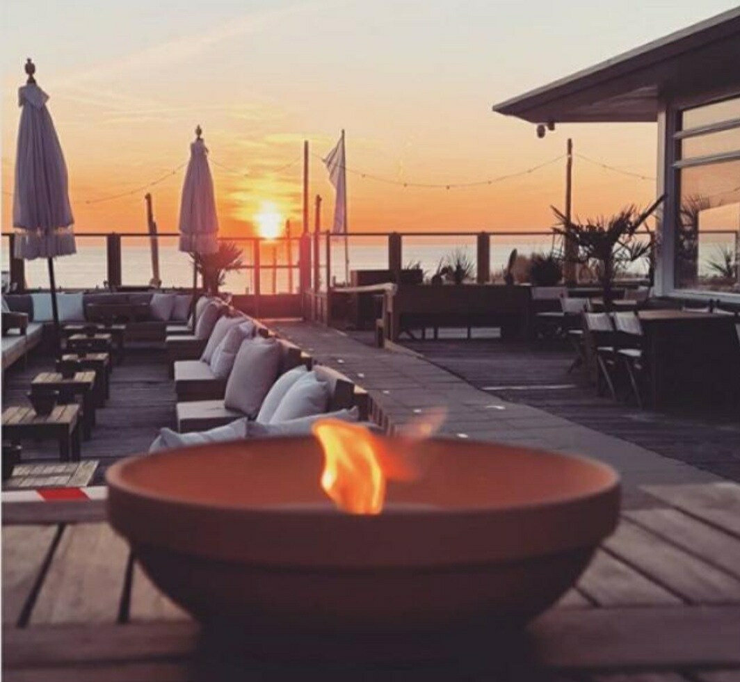 An outdoor terrace is captured at sunset, featuring a cozy fire bowl at the forefront. Comfortable seating areas with plush cushions are arranged along the deck, and the ocean stretches out in the background, reflecting the warm glow of the evening sky.