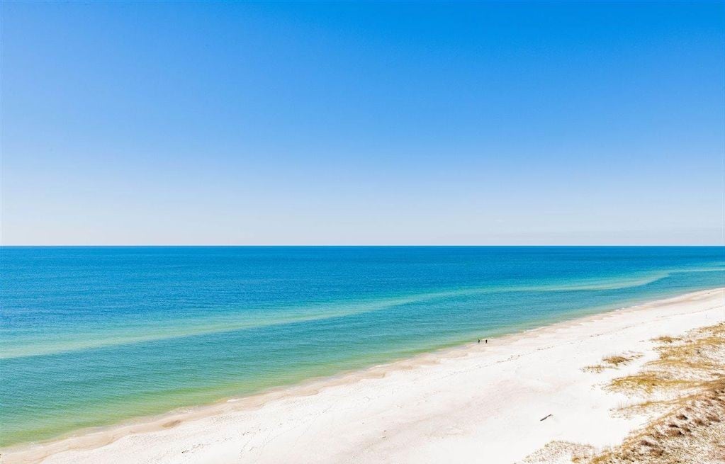 A serene view of the Gulf of Mexico is captured, showcasing tranquil turquoise waters meeting a soft, sandy beach. The horizon is clear, blending seamlessly with a bright blue sky, creating a sense of openness and tranquility.