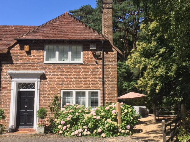 A charming brick house stands prominently against a clear blue sky. Lush hydrangea blooms frame the walkway, and a shaded patio area with a large umbrella is visible in the garden. The surrounding landscape includes tall trees, enhancing the property’s inviting appeal.