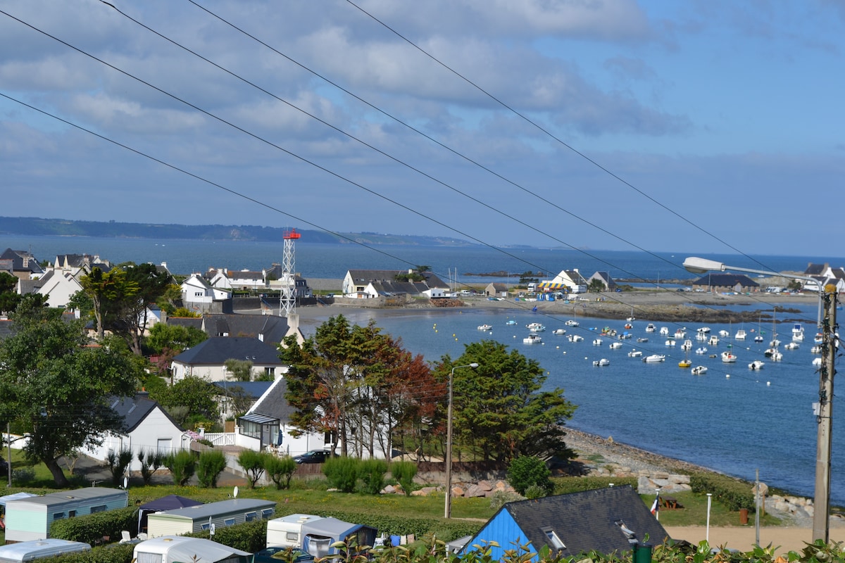 A coastal view showcases boats gently bobbing in the water, framed by residential buildings and greenery. The shoreline features a mix of homes and recreational areas, with distant hills visible against a partly cloudy sky, creating a serene atmosphere near the beach.