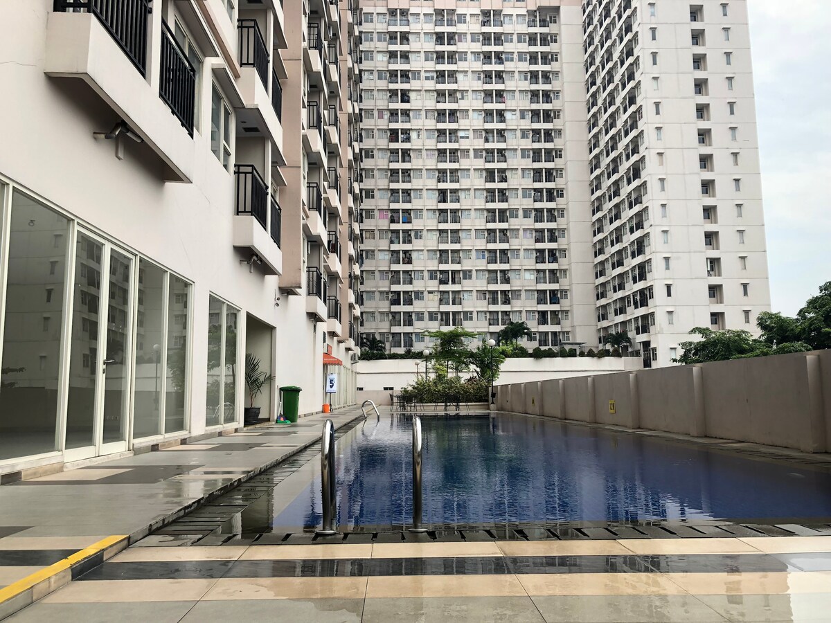 A serene swimming pool reflects light, bordered by modern apartment buildings. The well-maintained area is paved with tiles, providing ample space for relaxation. Glass doors lead to the pool area, offering a view of the lush surroundings.