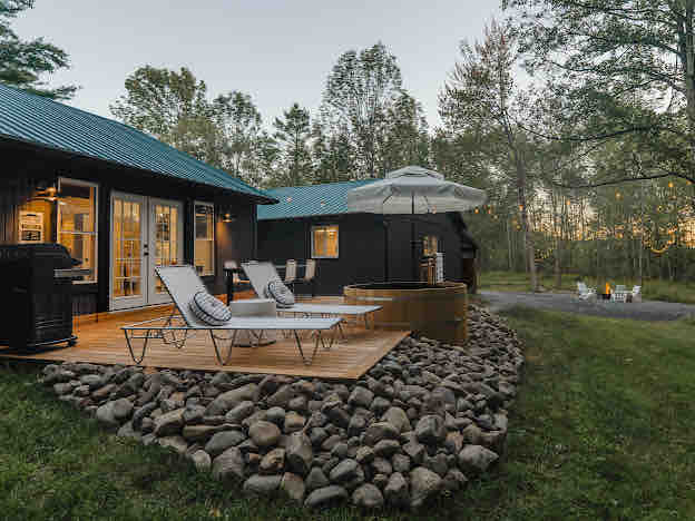 A private outdoor space features a wooden deck with reclining lounge chairs and a large umbrella. A cedar hot tub is positioned nearby, surrounded by smooth stones. In the background, the natural landscape includes trees and a fire pit area with chairs, indicating a serene environment.
