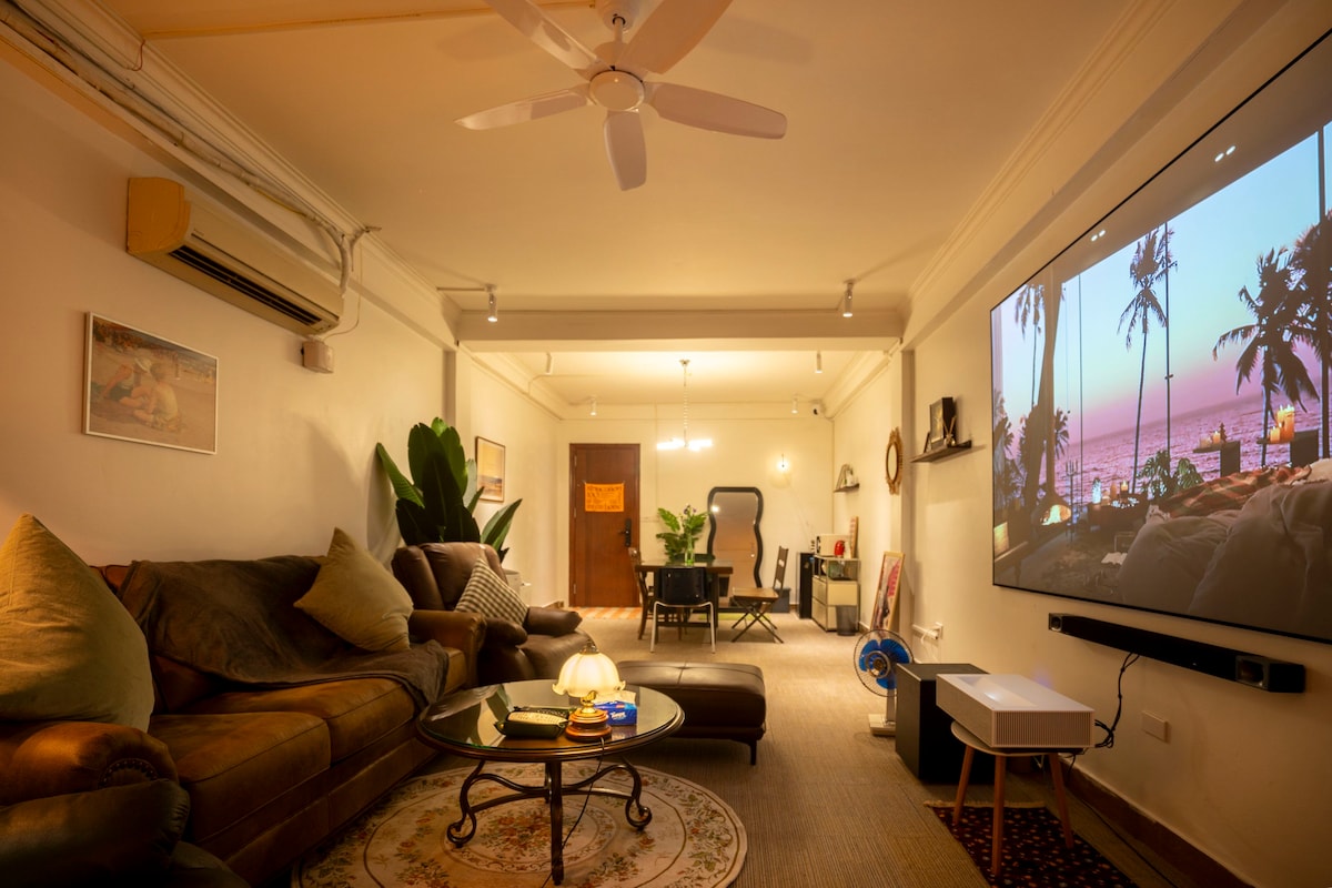 A cozy room is shown with a large projection screen displaying a serene sunset scene. A comfortable brown sofa is complemented by decorative cushions. A circular rug sits beneath a coffee table, and various plants are placed throughout the space, contributing to a warm atmosphere.
