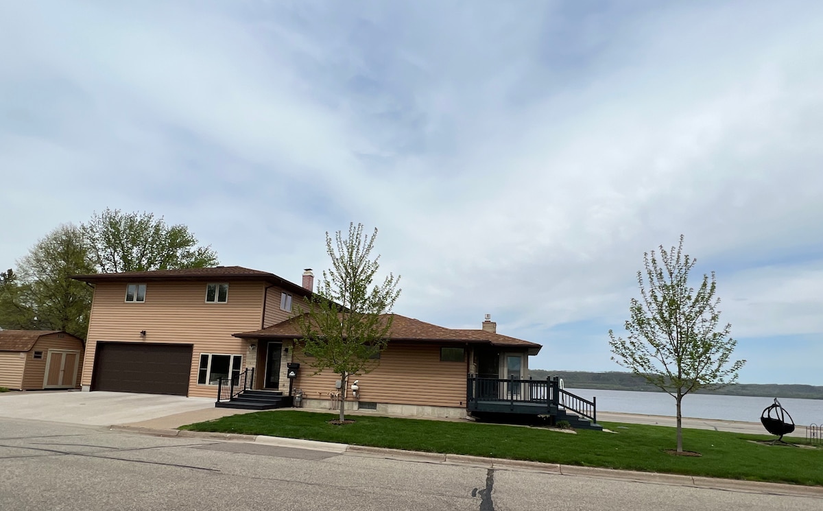 A spacious two-story house is shown, with a light brown exterior and a covered front porch. Lush green grass is seen in the front yard, alongside young trees. The property is situated near a serene body of water, with a cloudy sky above.