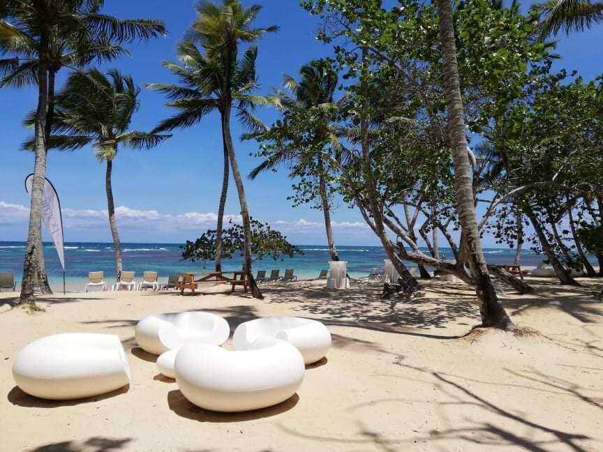 A sandy beach is adorned with modern white lounge seating under swaying palm trees. The turquoise water glistens in the distance, and a few shaded areas are visible among the trees. White chairs are arranged in the background, suggesting a tranquil gathering space.