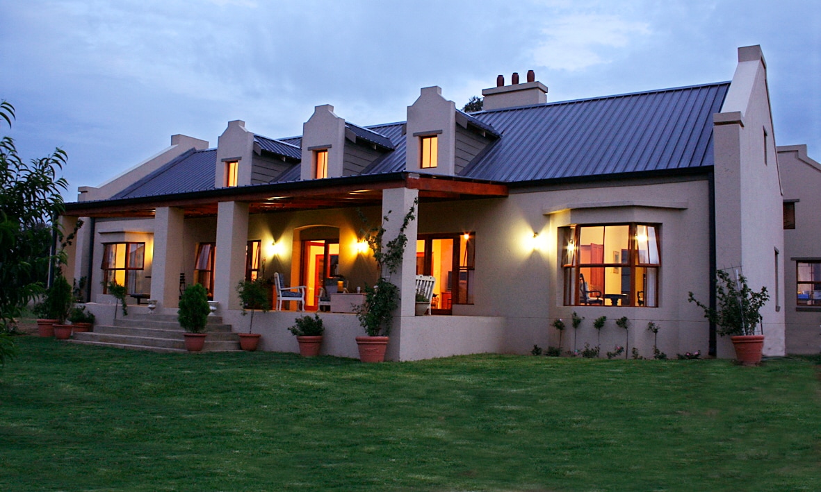 The exterior of a spacious retreat is showcased, featuring a covered porch with seating areas. Large windows illuminate the façade, reflecting a warm glow in the evening light. Surrounding greenery and potted plants add to the inviting outdoor space.