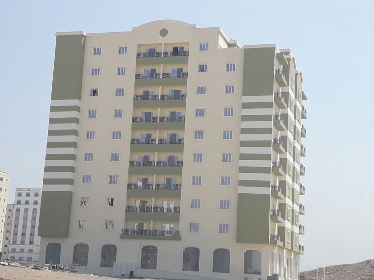 A modern, six-story building features a clean facade with light yellow and olive green accents. Multiple balconies are visible on each floor, showcasing a uniform design. Large windows allow ample natural light, and the ground floor includes an entryway with visible openings.