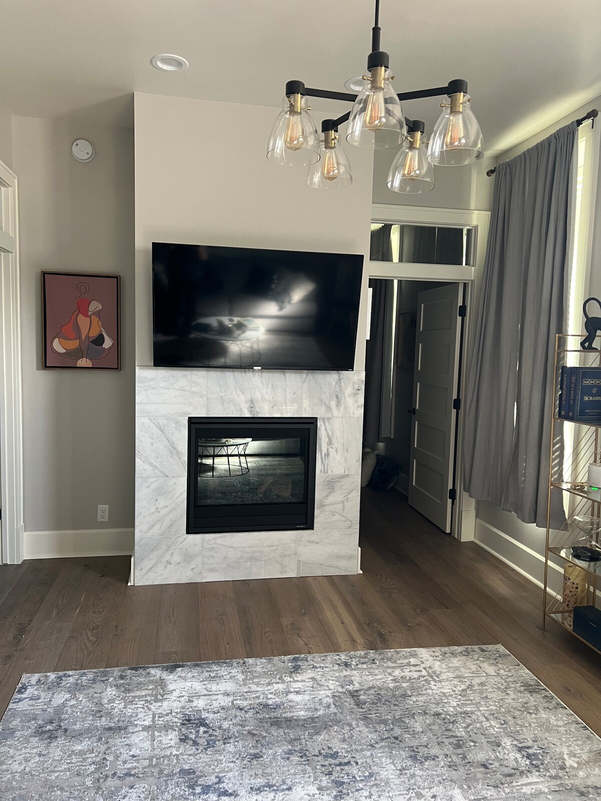 A living area features a modern fireplace framed in marble beneath a mounted flat-screen television. Light fixtures with exposed bulbs hang from the ceiling, while soft gray curtains drape beside the window. A patterned area rug adds texture to the warm wooden floor.