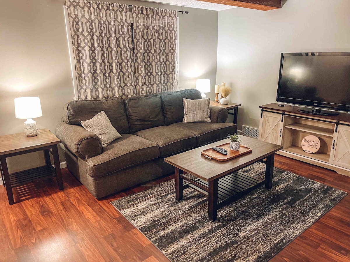 A comfortable living area is highlighted by a spacious dark grey couch and a wooden coffee table. Two lamps provide soft lighting, while a patterned curtain partially covers a window. A flat-screen TV is positioned against the wall, contributing to the inviting atmosphere.
