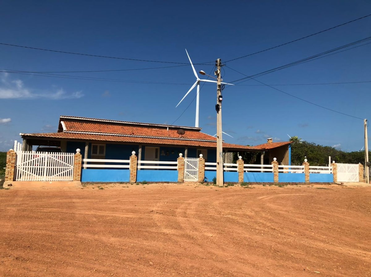 A traditional house is displayed with a vibrant blue exterior and a terracotta roof. A white fence encircles the property, complemented by a wind turbine in the background. The surrounding area features a dirt road highlighting the house's peaceful rural setting.
