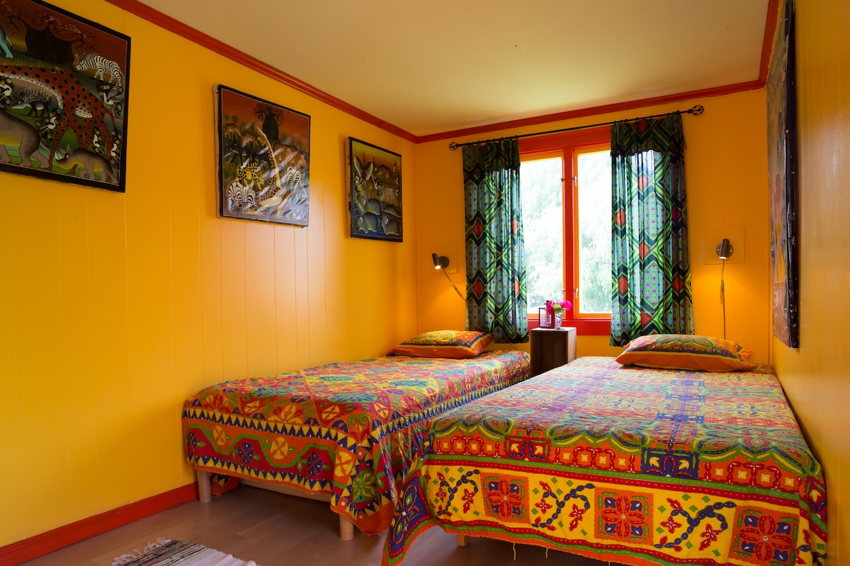 The room features two beds adorned with vibrant patterned bedspreads. Bright yellow walls create a cheerful atmosphere, complemented by colorful artwork. Natural light enters through large windows, dressed with matching curtains. A small bedside table holds a kettle, enhancing the room's functionality.