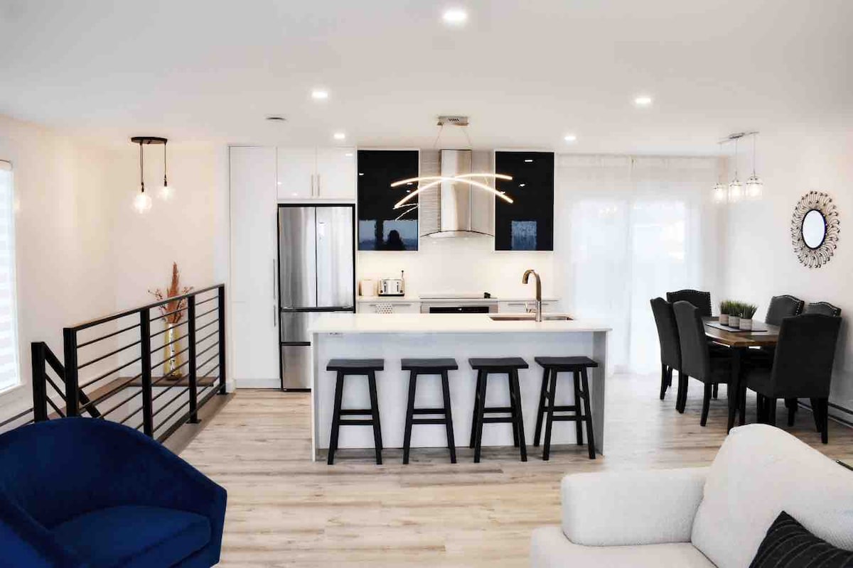 The spacious kitchen and dining area feature a contemporary design with a large island and modern appliances. Bright lighting illuminates the open space, complemented by a dining table and comfortable seating. Large windows allow natural light to flow in, enhancing the inviting atmosphere.