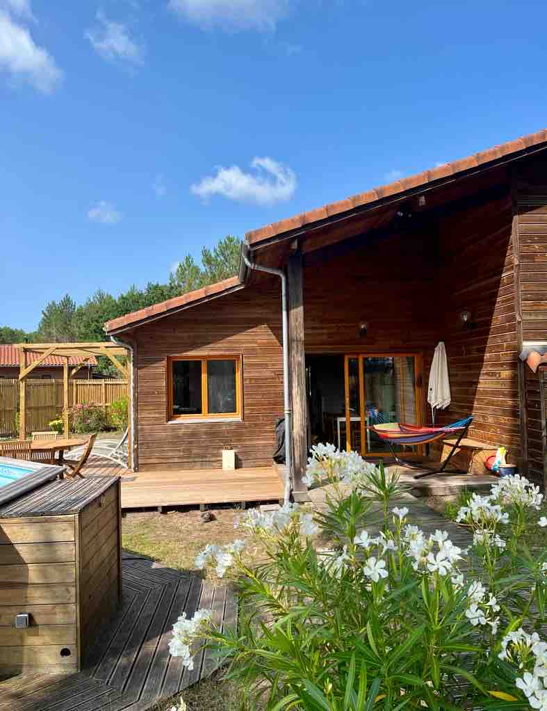 A wooden exterior of the ecological house is shown, featuring a spacious deck with a hammock and a view of nearby greenery. A private pool is visible, alongside blooming white flowers in the garden, under a bright blue sky with scattered clouds.