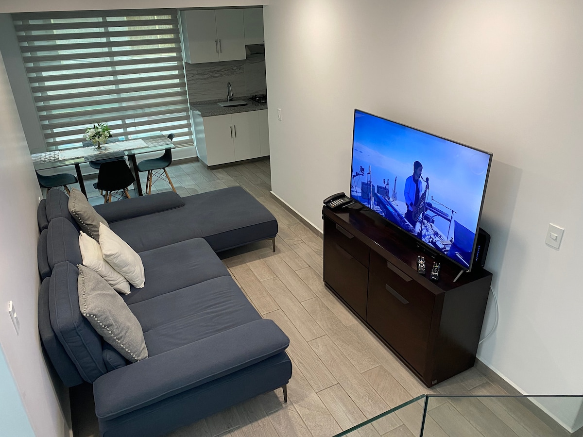 A modern living area features a spacious sectional sofa in navy blue, complemented by soft pillows. A large flat-screen television is positioned on a dark wooden cabinet. An adjacent dining area is visible, equipped with a table and chairs, with natural light filtering through window treatments.