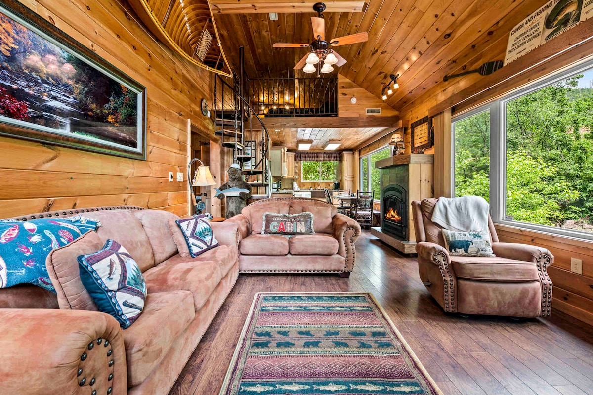 The living room features a spacious seating area with a plush sofa and armchair, accented by a rustic area rug. Large windows offer views of the surrounding greenery, while the indoor fireplace and wooden ceiling add cozy charm. A spiral staircase leads to the loft area.