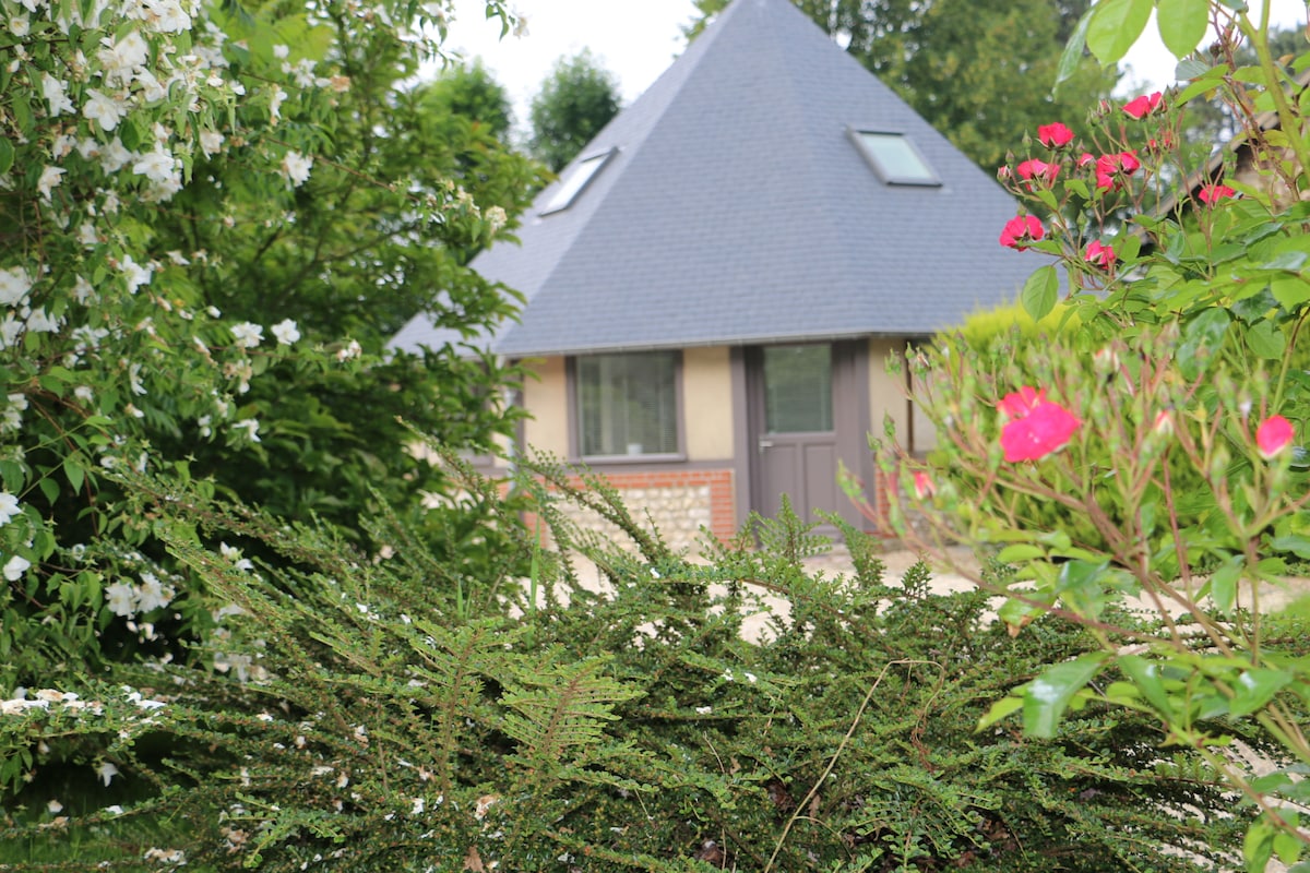 A charming house with a distinctive triangular roof is framed by lush greenery and colorful flowers. The entrance features large windows, and the surrounding foliage creates a private ambiance, showcasing the serene garden environment.