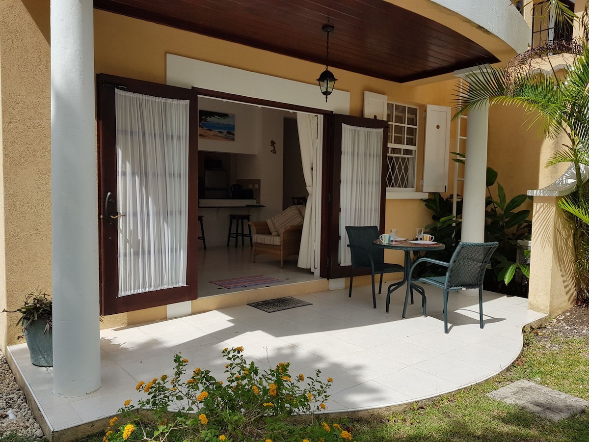 A covered patio is seen, featuring a small round table with two chairs. Glass doors lead into the living area, surrounded by tropical plants. The floor is tiled in light colors, enhancing the openness with a view of the indoor space.