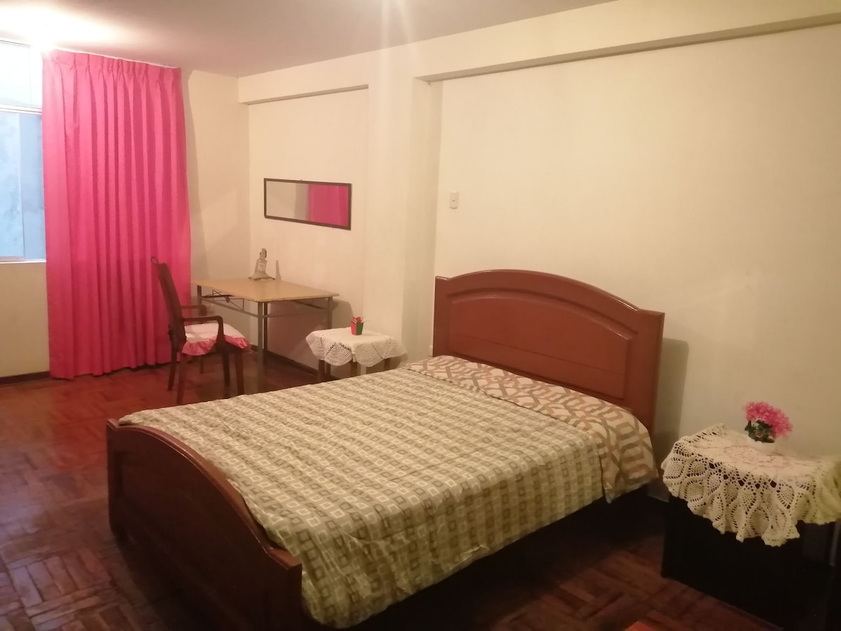 A spacious bedroom features a large bed with a patterned comforter, complemented by a bedside table adorned with a small bouquet of flowers. A wooden desk and chair are offered for work, with a mirror positioned above the desk. Natural light enters through a window, with pink curtains adding color.