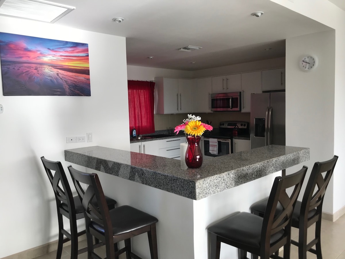 A modern kitchen area is shown with a granite countertop and seating for four on dark wooden stools. A colorful landscape artwork adorns the wall above the counter. The kitchen features stainless steel appliances and white cabinetry, with natural light filtering through a nearby window covered by red drapes.