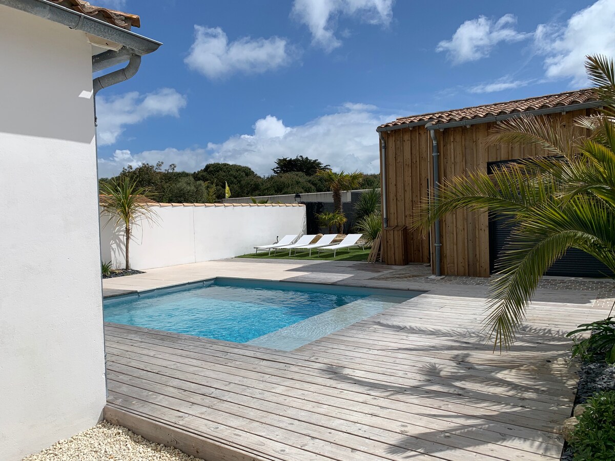 A swimming pool is surrounded by a wooden deck and lounge chairs under a bright sky with scattered clouds. Nearby, palm trees and a wooden structure enhance the outdoor area, with a wall providing privacy.