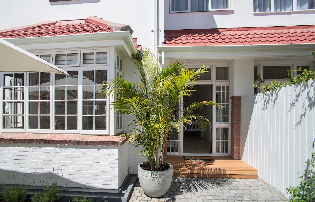 The entrance area is framed by large windows that allow natural light to flow into the space. A potted palm tree adds greenery, while the wooden steps lead to an inviting doorway. The quaint courtyard features cobblestone paving and a white fence for added privacy.