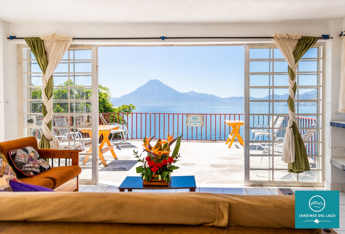 A bright living area features large glass doors that open to a private balcony overlooking Lake Atitlán and its surrounding volcanoes. Comfortable seating is arranged to take advantage of the scenic views, while lush greenery and a colorful arrangement of flowers add a touch of nature.