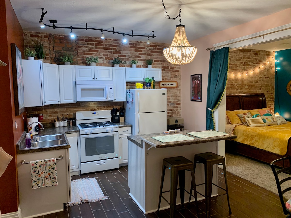 A modern kitchen area features white cabinetry and stainless steel appliances, with a brick accent wall adding texture. A round light fixture hangs above a central island with bar stools. The living space includes a warm color scheme and decorative curtains, leading to a cozy sleeping area.
