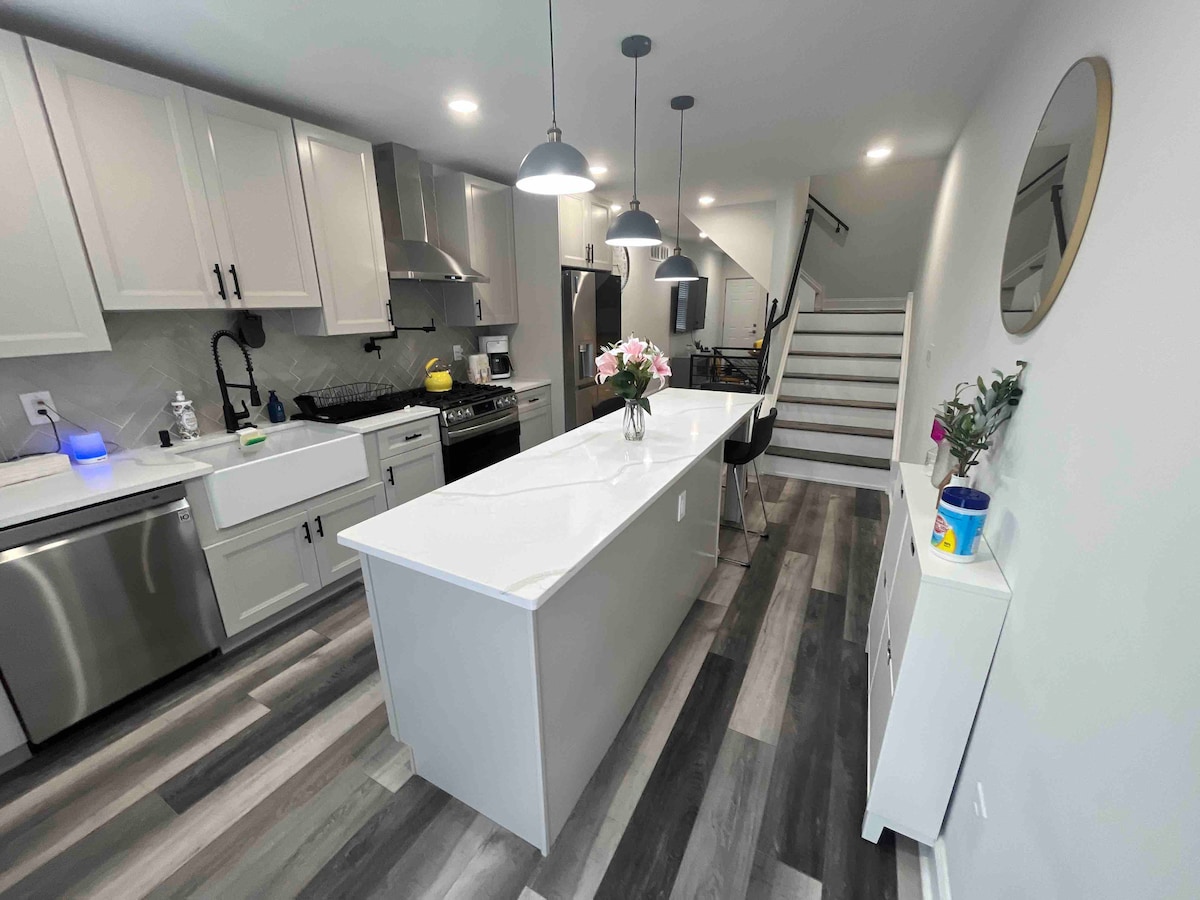 The modern kitchen features an island with a white countertop and seating for three. Stainless steel appliances, including a gas stove, are complemented by sleek cabinets. Natural light fills the space, highlighting the laminate flooring and creating a clean, inviting environment.
