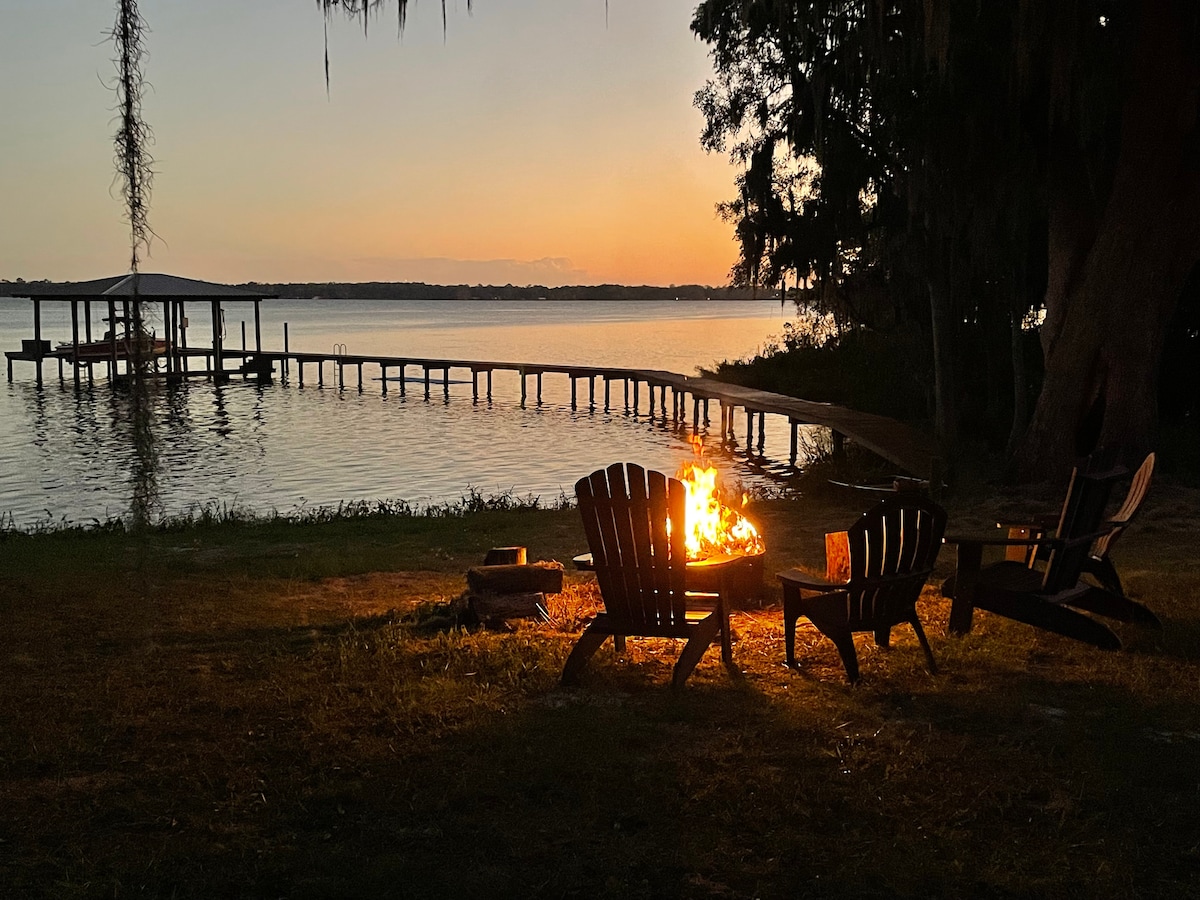 A cozy campfire is surrounded by several Adirondack chairs, set against a serene river view at sunset. A wooden dock extends into the water, framed by trees draped with Spanish moss, creating a peaceful and tranquil outdoor setting.